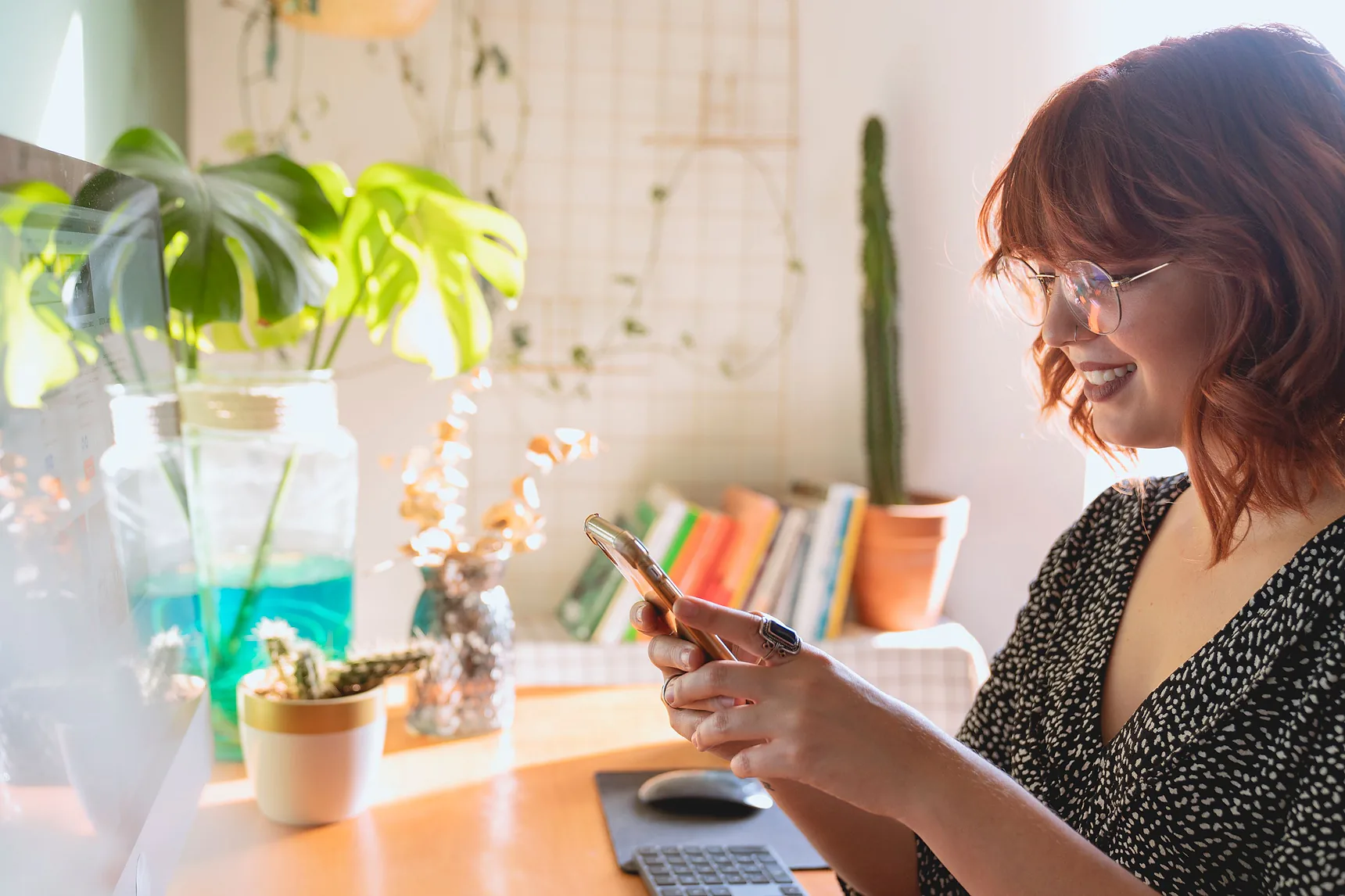 Smiling woman typing on her phone at a desk with plants and books behind