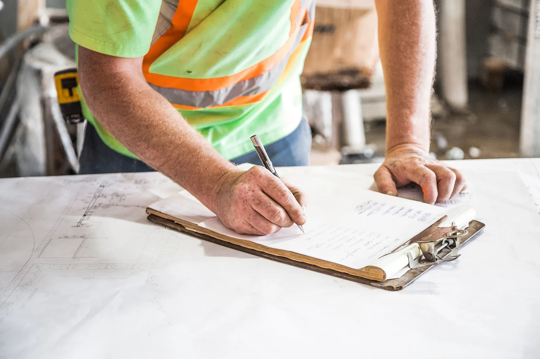 Worker in hi-vis filling in a checklist on a clipboard over plans