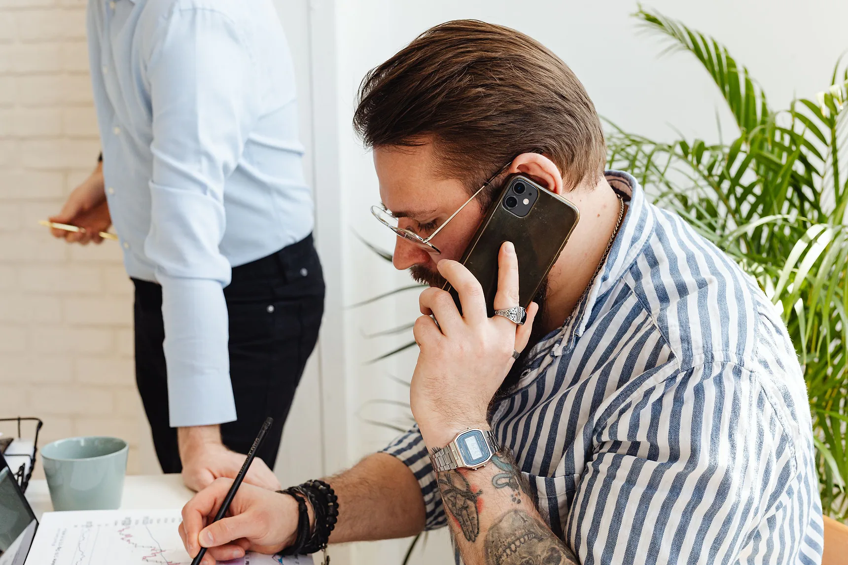 Man on a phone call writing notes at a desk while a colleague stands beside him